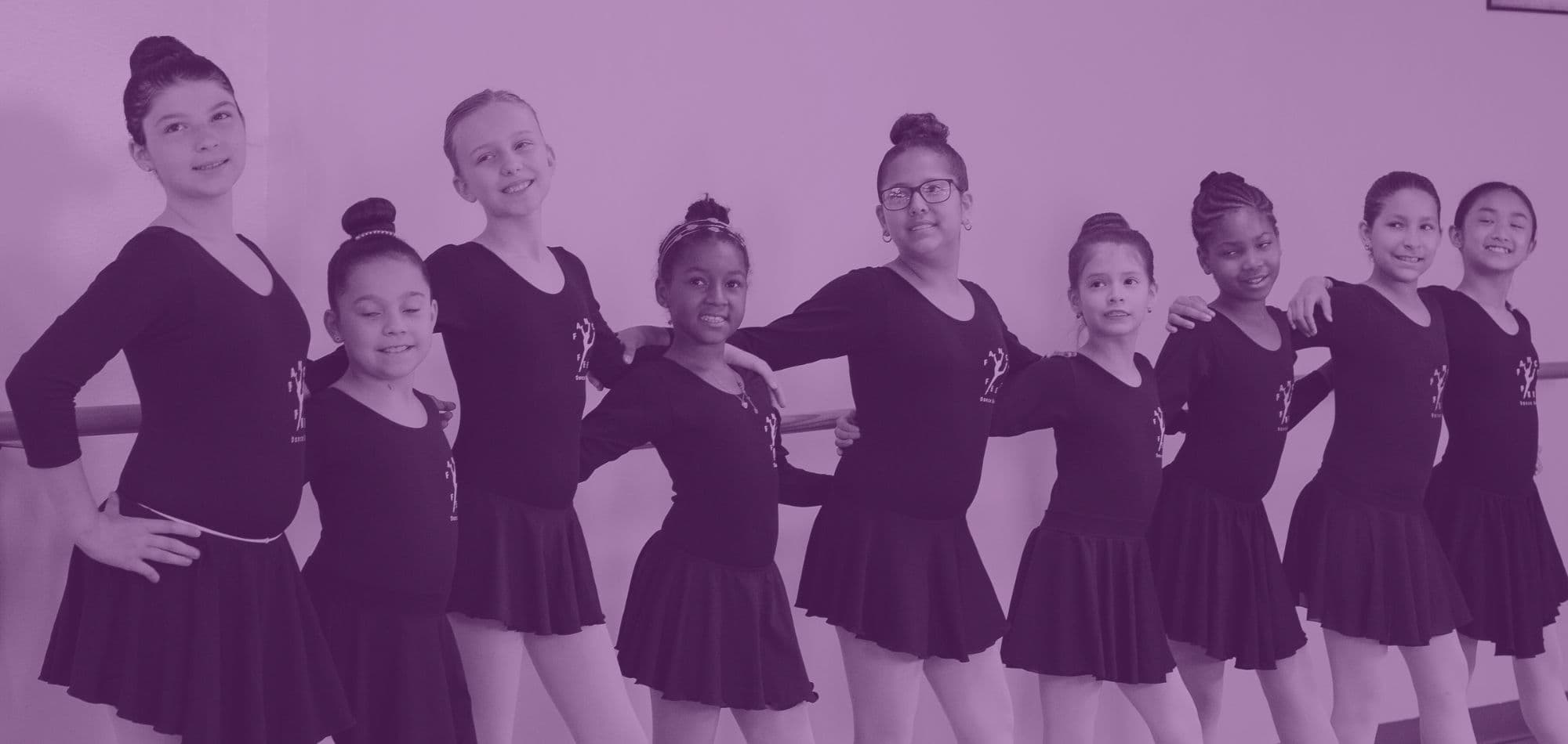 A line of Fancy Feet ballet students in black leotards smiling at the barre